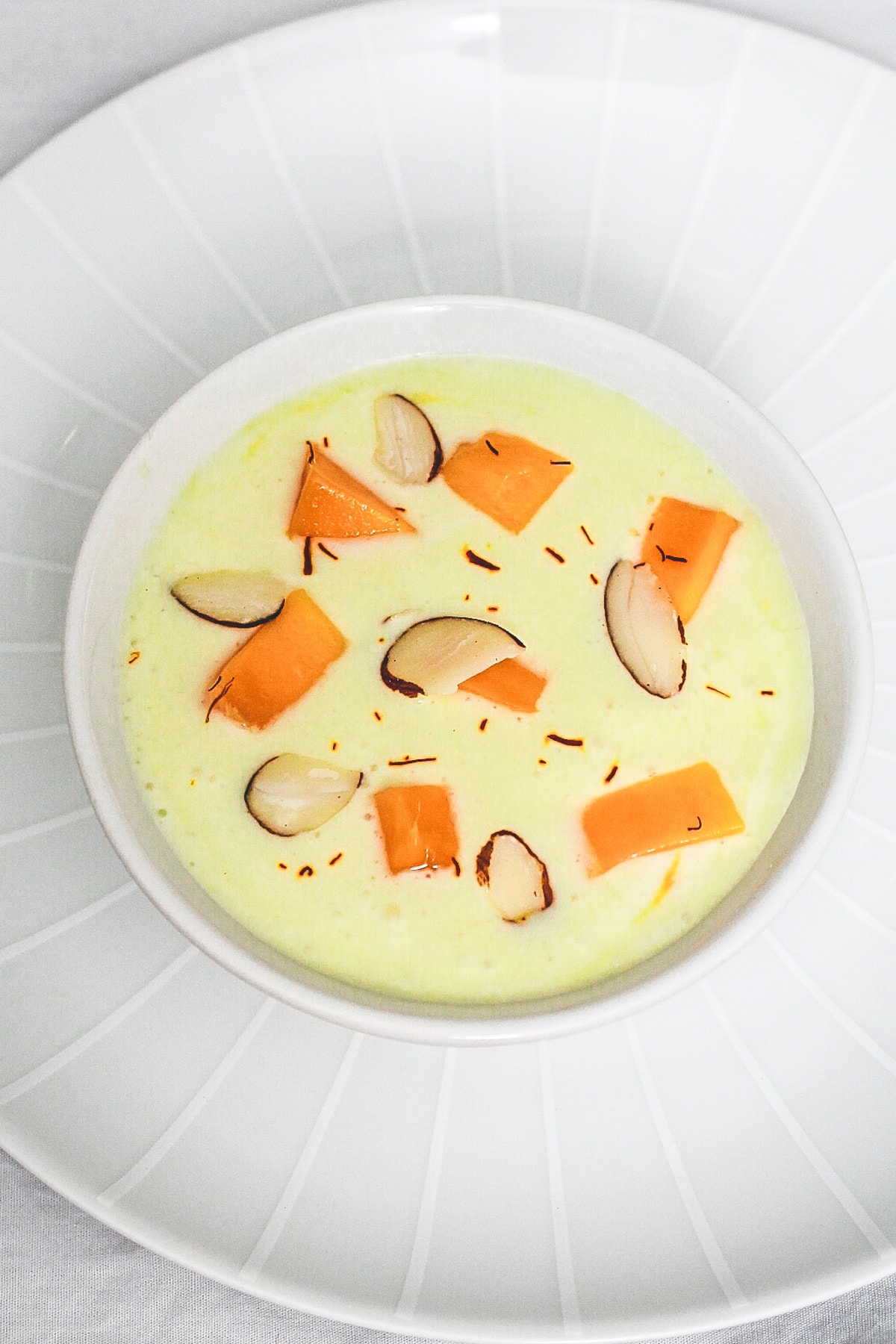 Mango raita served in a bowl and plate underneath, raita garnished with almonds, mango and saffron.