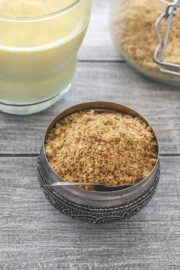 Milk masala powder with a spoon in it with masala doodh glass and a container in the back.