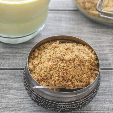 Milk masala powder with a spoon in it with masala doodh glass and a container in the back.
