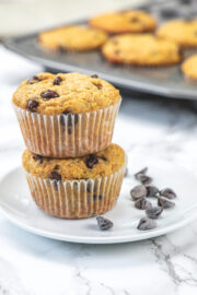 2 orange muffins are stacked in a small plate with chocolate chips on side and muffin tray in back.