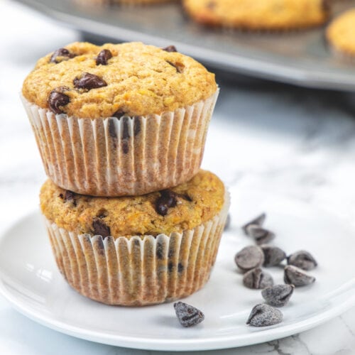 2 orange muffins are stacked in a small plate with chocolate chips on side and muffin tray in back.