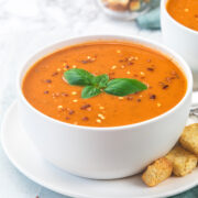 a bowl of roasted tomato soup garlished with basil and chili flakes with croutons on side.