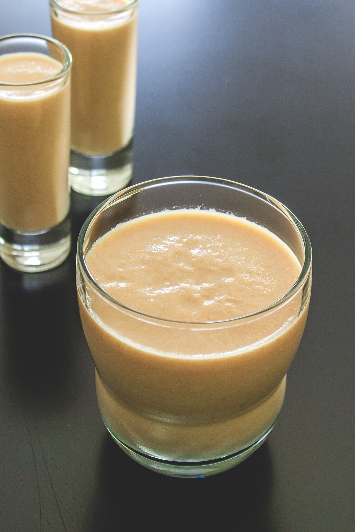 A glass of chikoo milkshake with 2 shot glasses in the back.