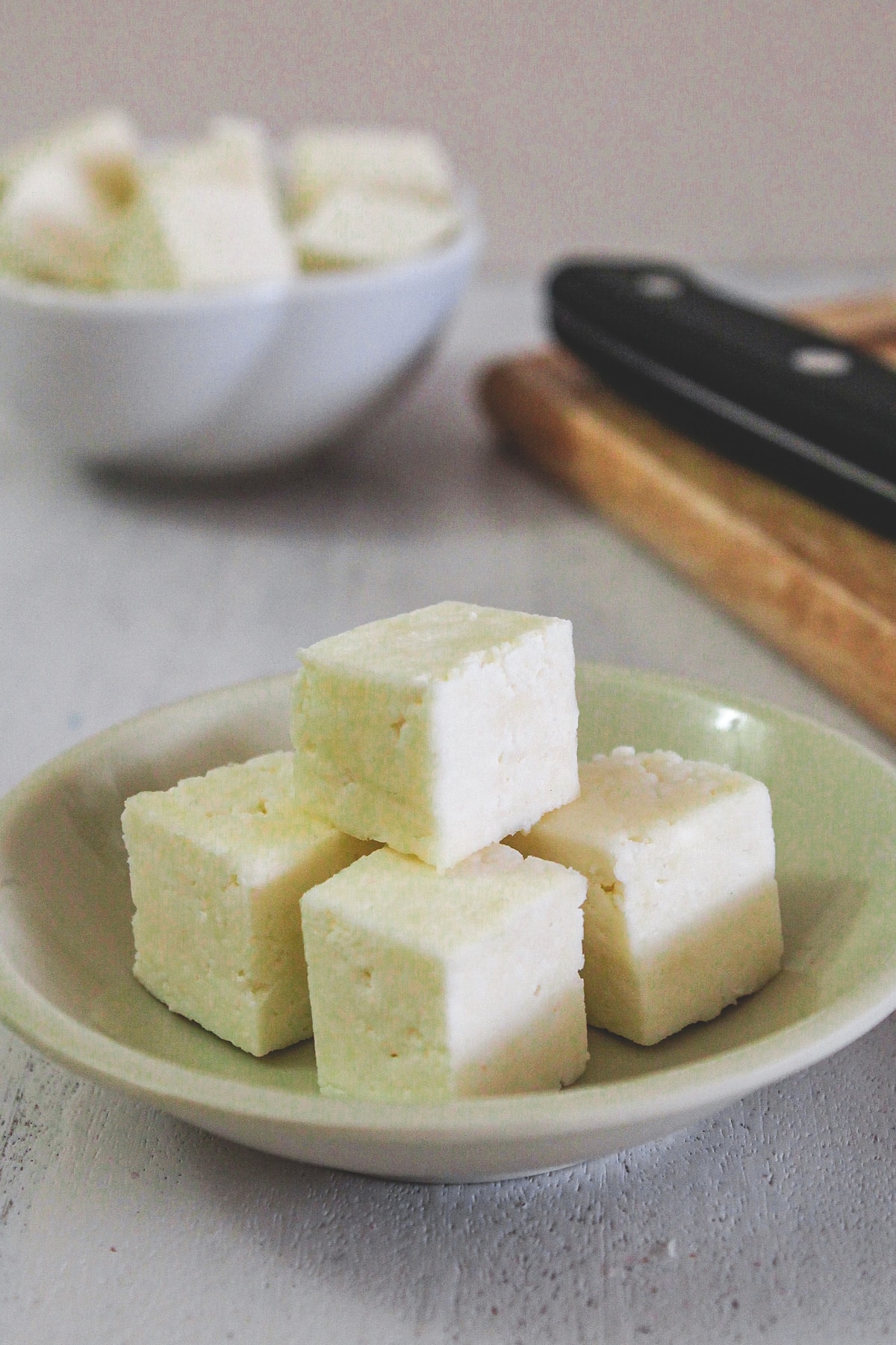 Paneer cubes in a small plate with knife and a bowl of paneer in the back.