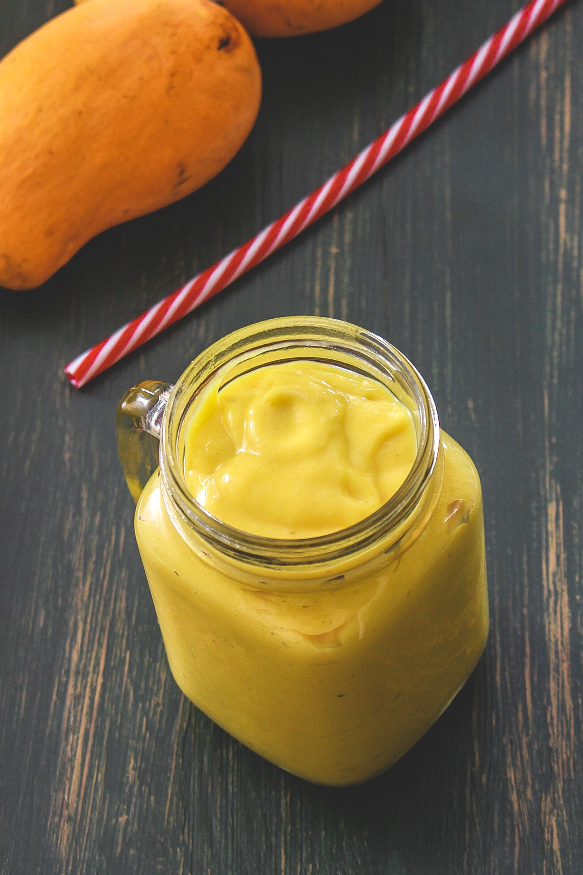 A jar of mango banana smoothie with straw and mangoes in the back side.