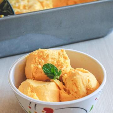 3 scoops of mango ice cream in a bowl and garnished with a mint leaf with loaf pan in back.
