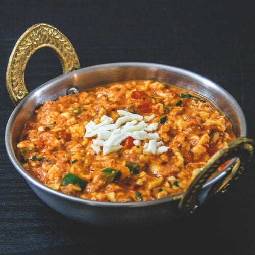 Paneer bhurji gravy in kadai serving bowl with raw grated paneer sprinkled on top.