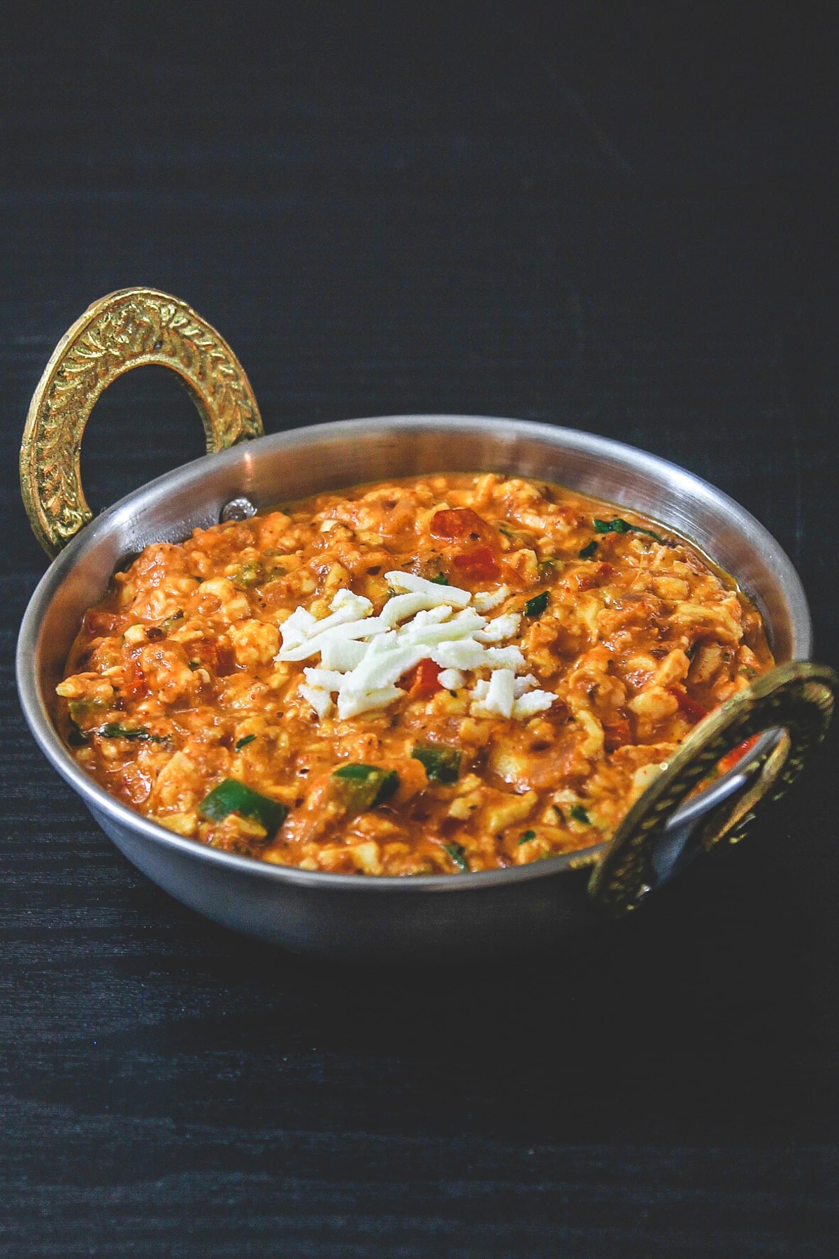 Paneer bhurji gravy in kadai serving bowl with raw grated paneer sprinkled on top.