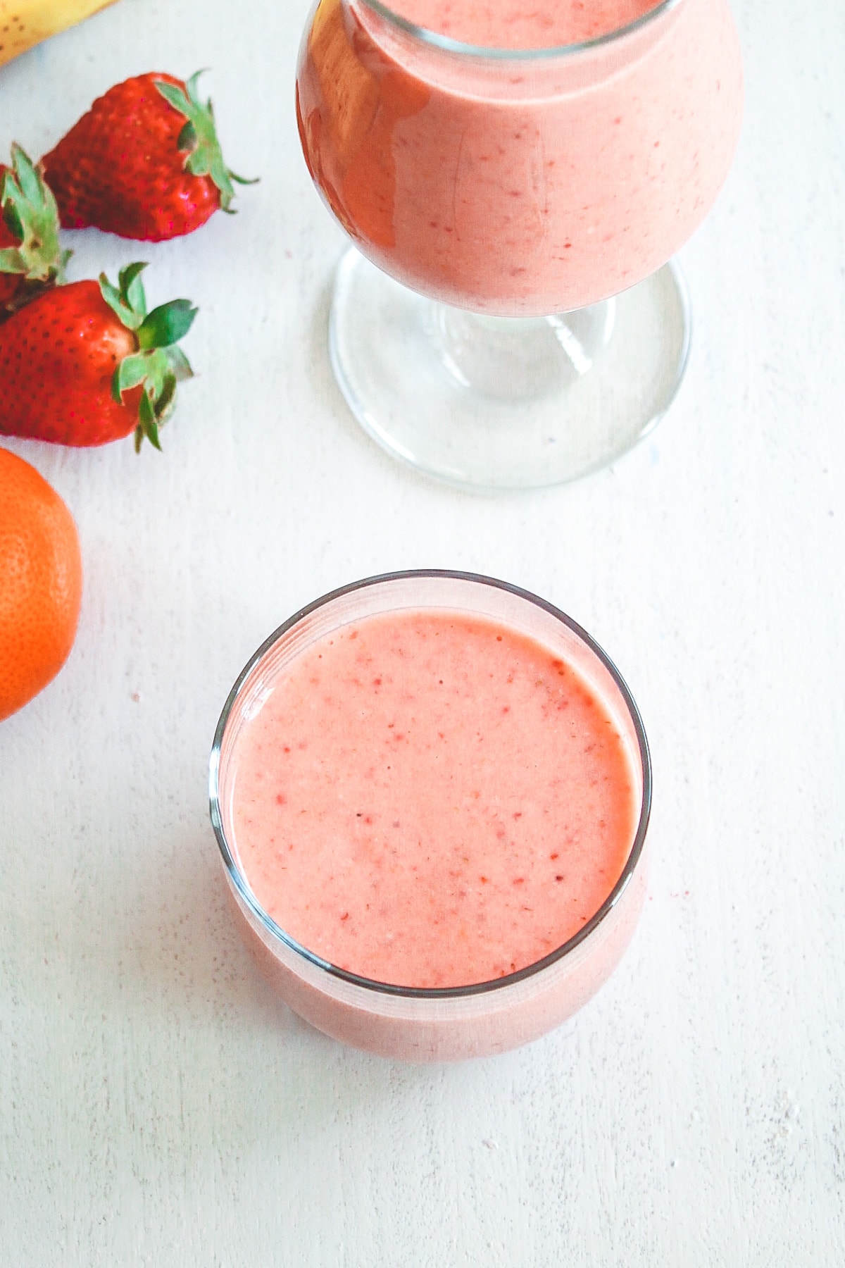 2 glasses of strawberry banana yogurt smoothie with strawberries and clementine in the back.