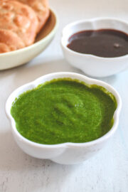 A bowl of green chutney with a bowl of tamarind date chutney and kachori in the back.