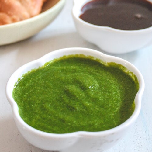 A bowl of green chutney with a bowl of tamarind date chutney and kachori in the back.