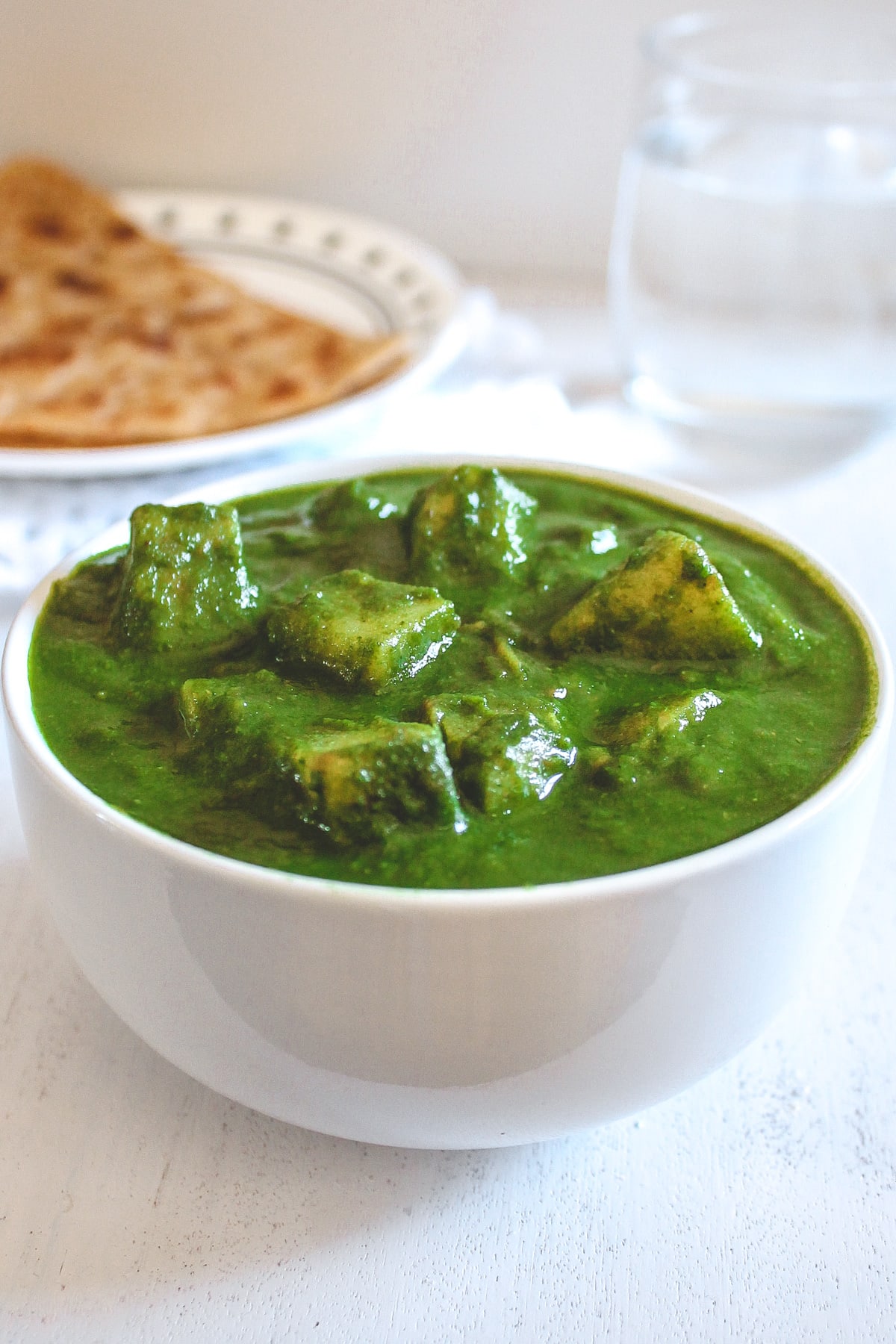 Aloo palak in a white bowl with paratha and glass of water in the back side.