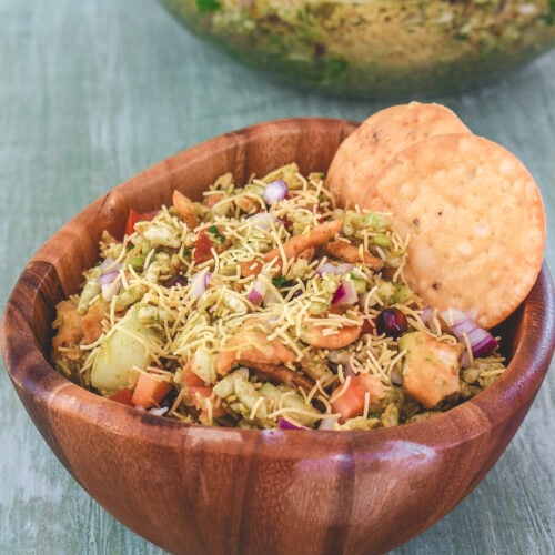 Bhel puri in a wooden bowl with 2 papdi on side and more bhel in the back.