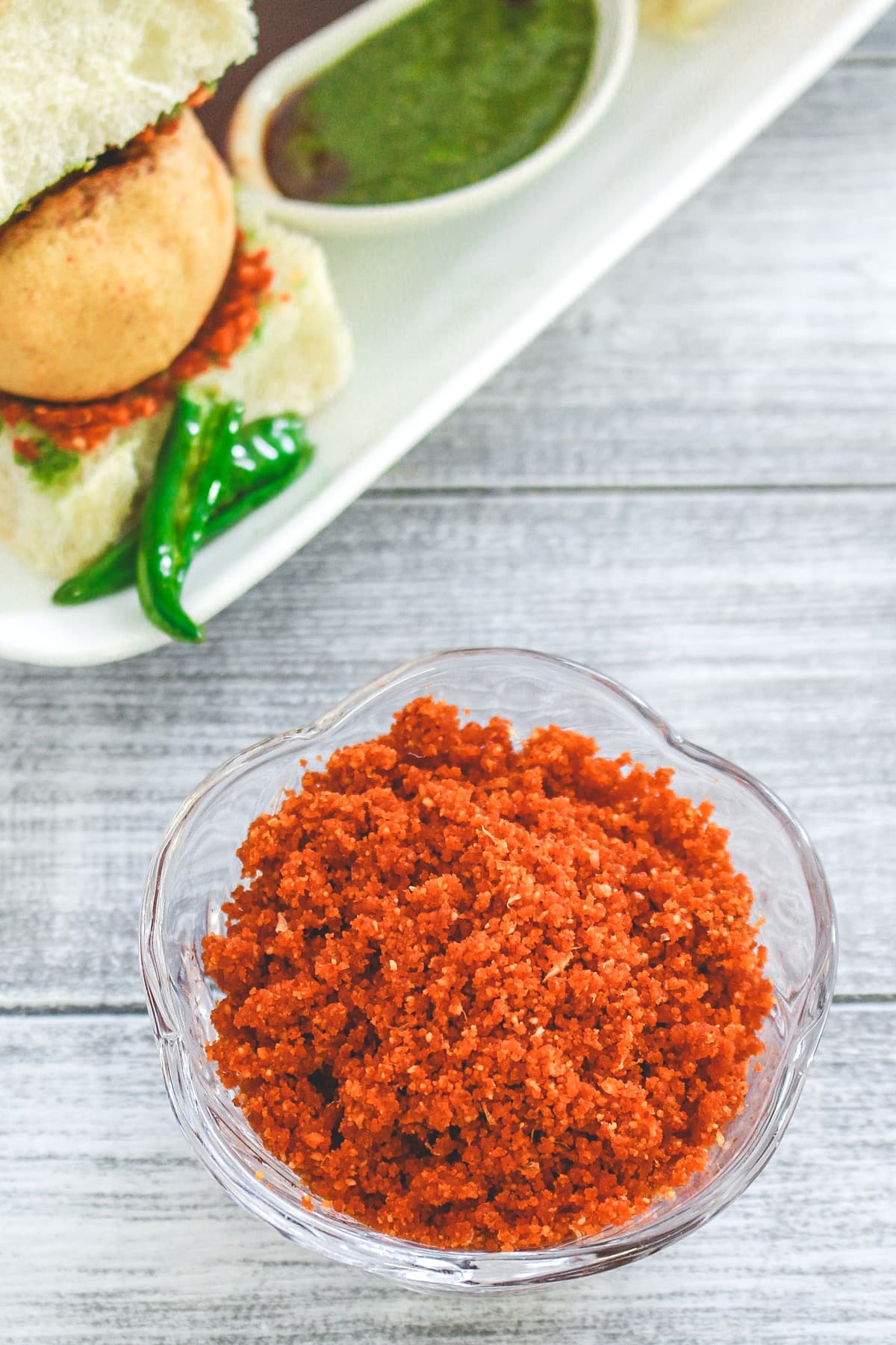 Dry garlic chutney in a bowl and vada pav with fried chilies in the back.