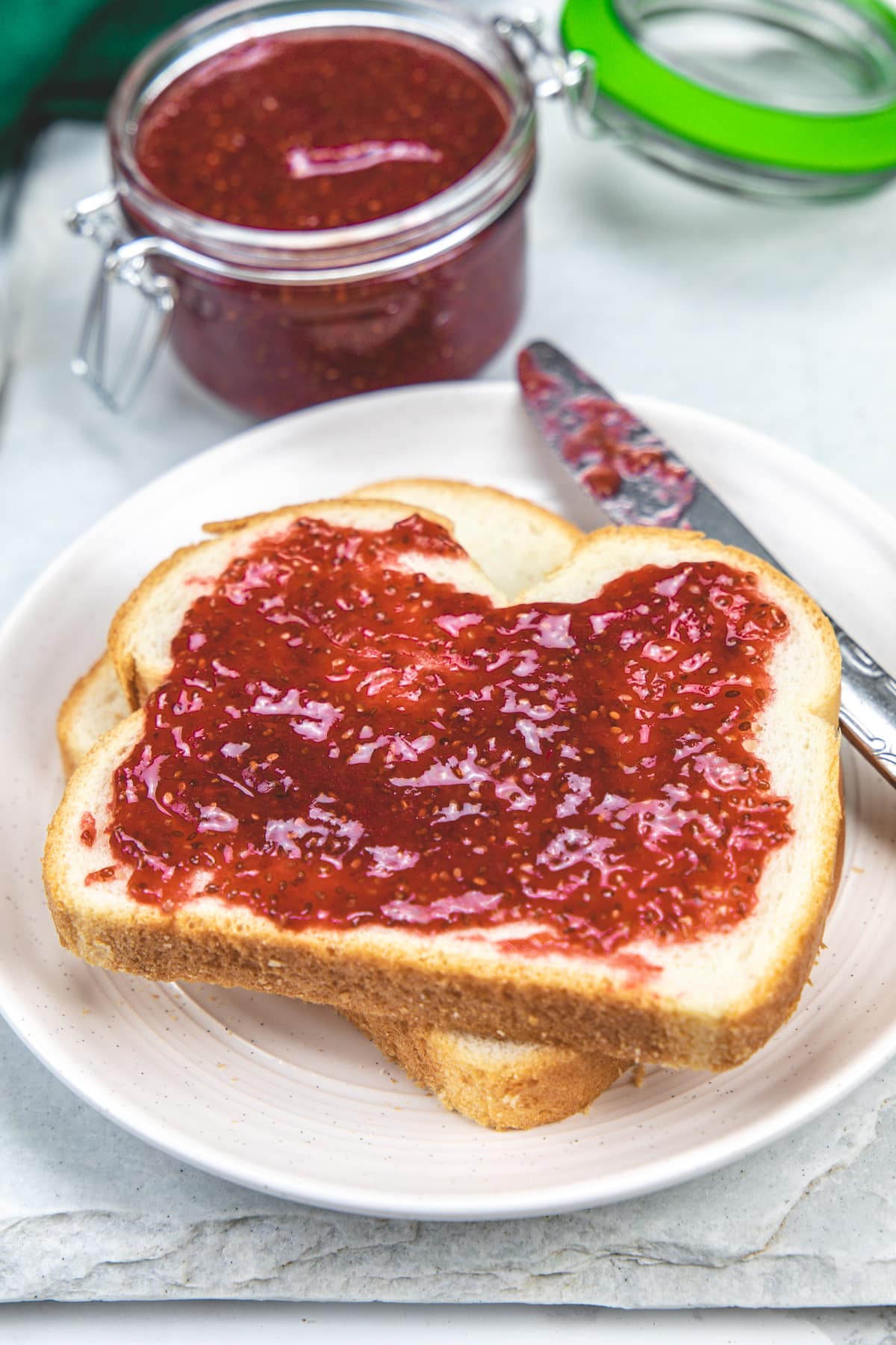 Strawberry chia jam spreaded on a bread slice with a jar of jam in the back.