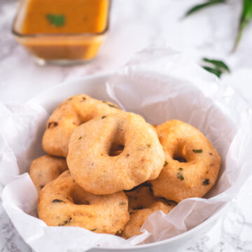 Medu vada in a bowl lined with parchment paper with sambar and chuteny in the back.