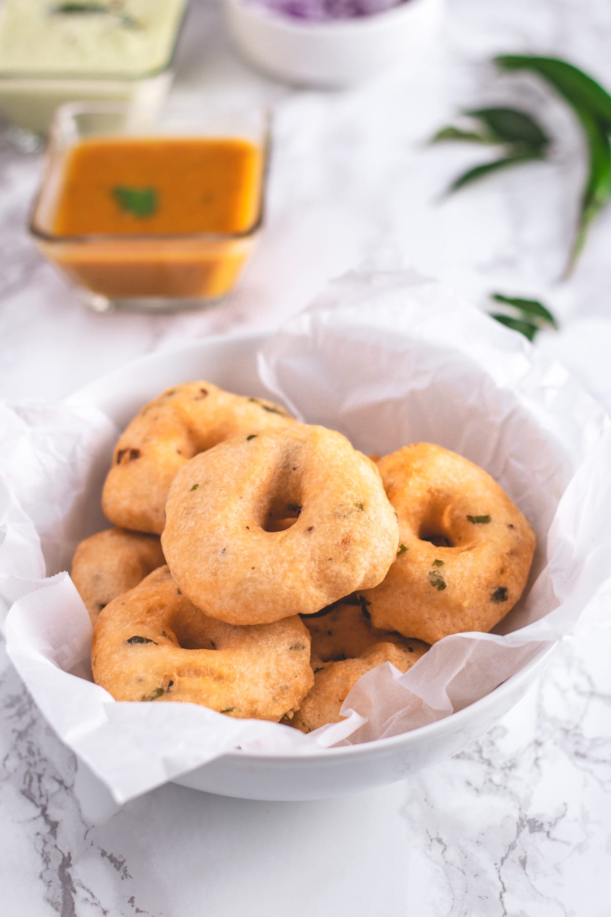 Medu vada in a bowl lined with parchment paper with sambar and chuteny in the back.