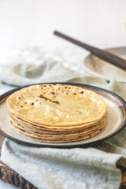 A stack of phulka roti in a plate with napkin under the plate and rolling pin in the back.