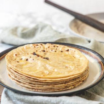 A stack of phulka roti in a plate with napkin under the plate and rolling pin in the back.