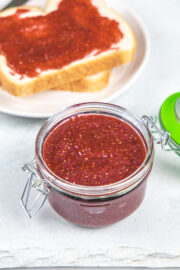 Strawberry chia jam in a glass jar with jam spreaded bread slice in the back.