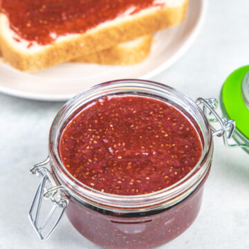 Strawberry chia jam in a glass jar with jam spreaded bread slice in the back.