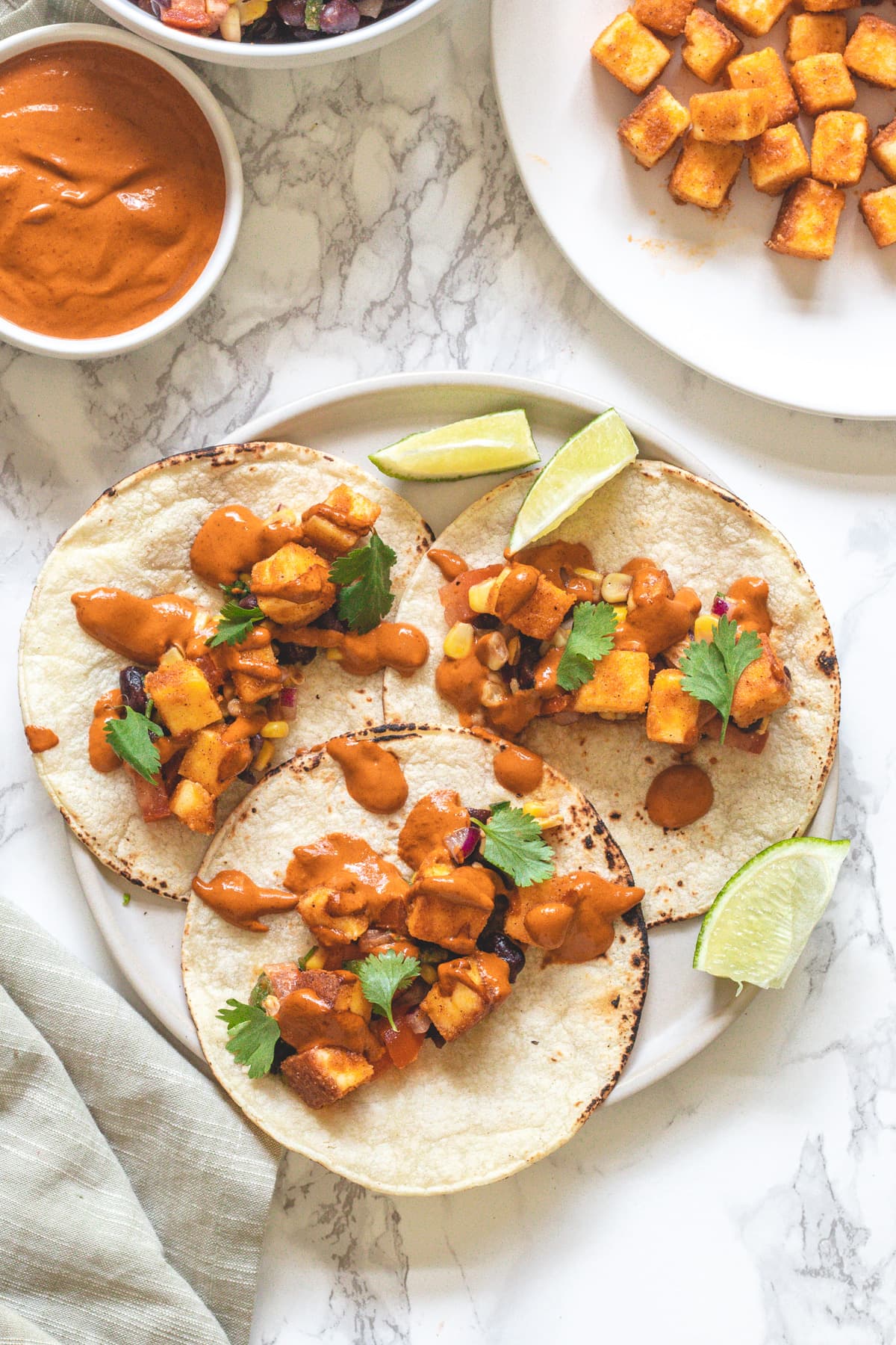 3 Paneer tacos in a plate with lime wedges, chipotle sauce and paneer on the side.
