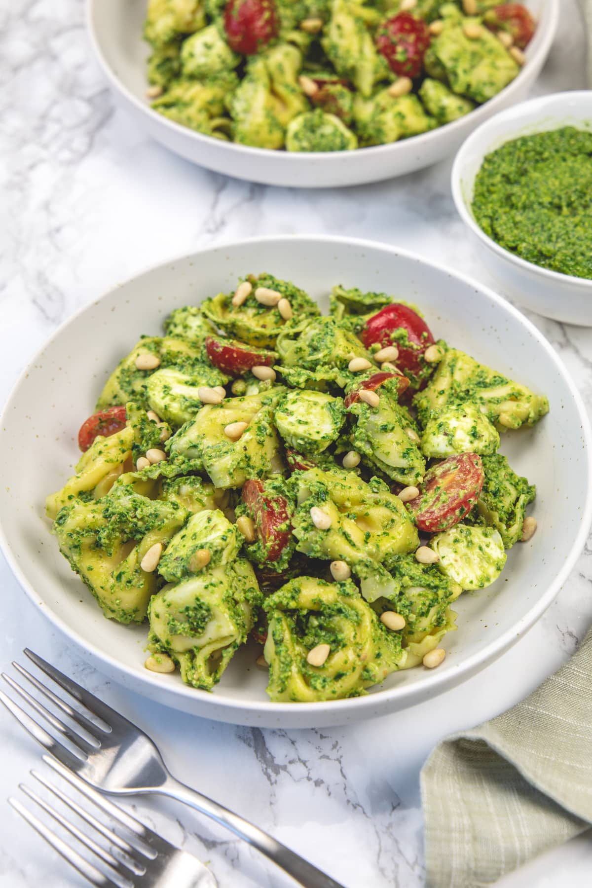 Pesto tortellini salad in two plates with forks on left and a bowl of extra pesto on right.