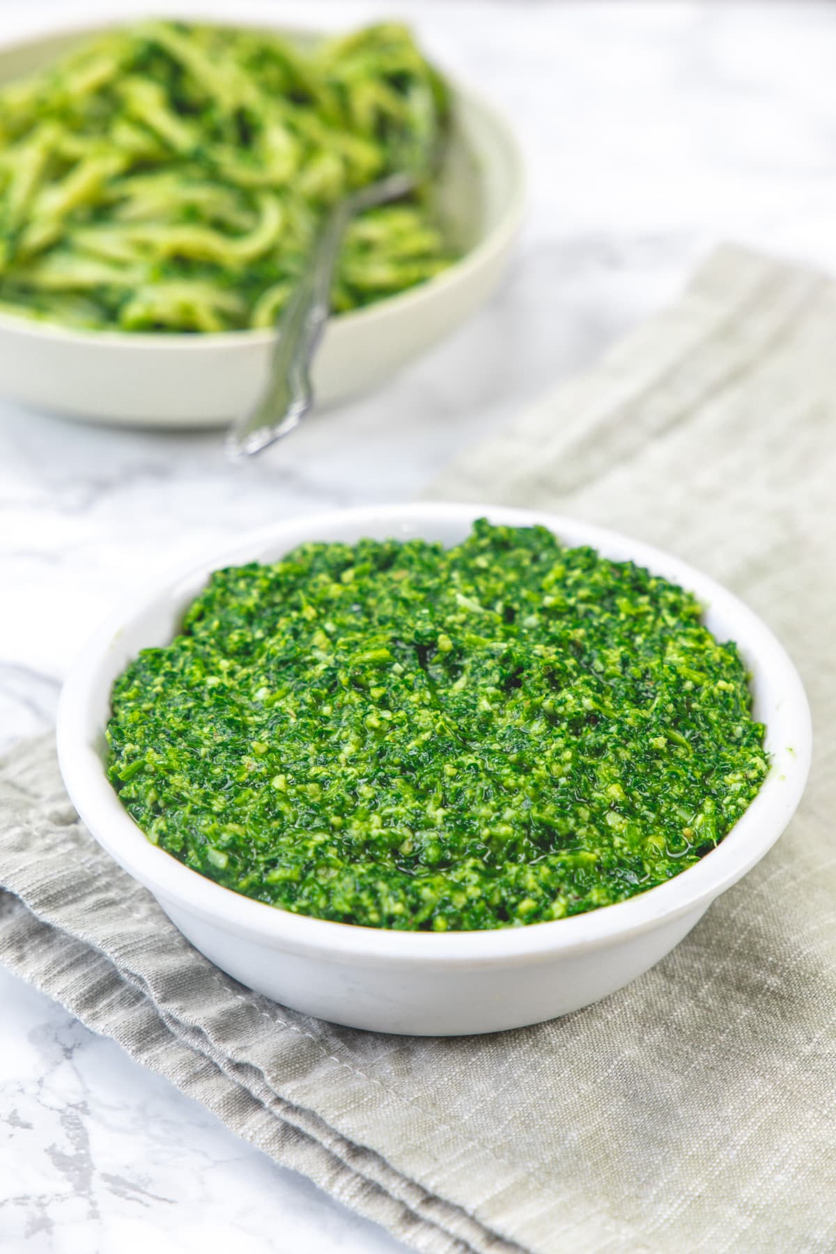 Parsley pesto in a bowl with napkin underneath and pasta plate in the back.