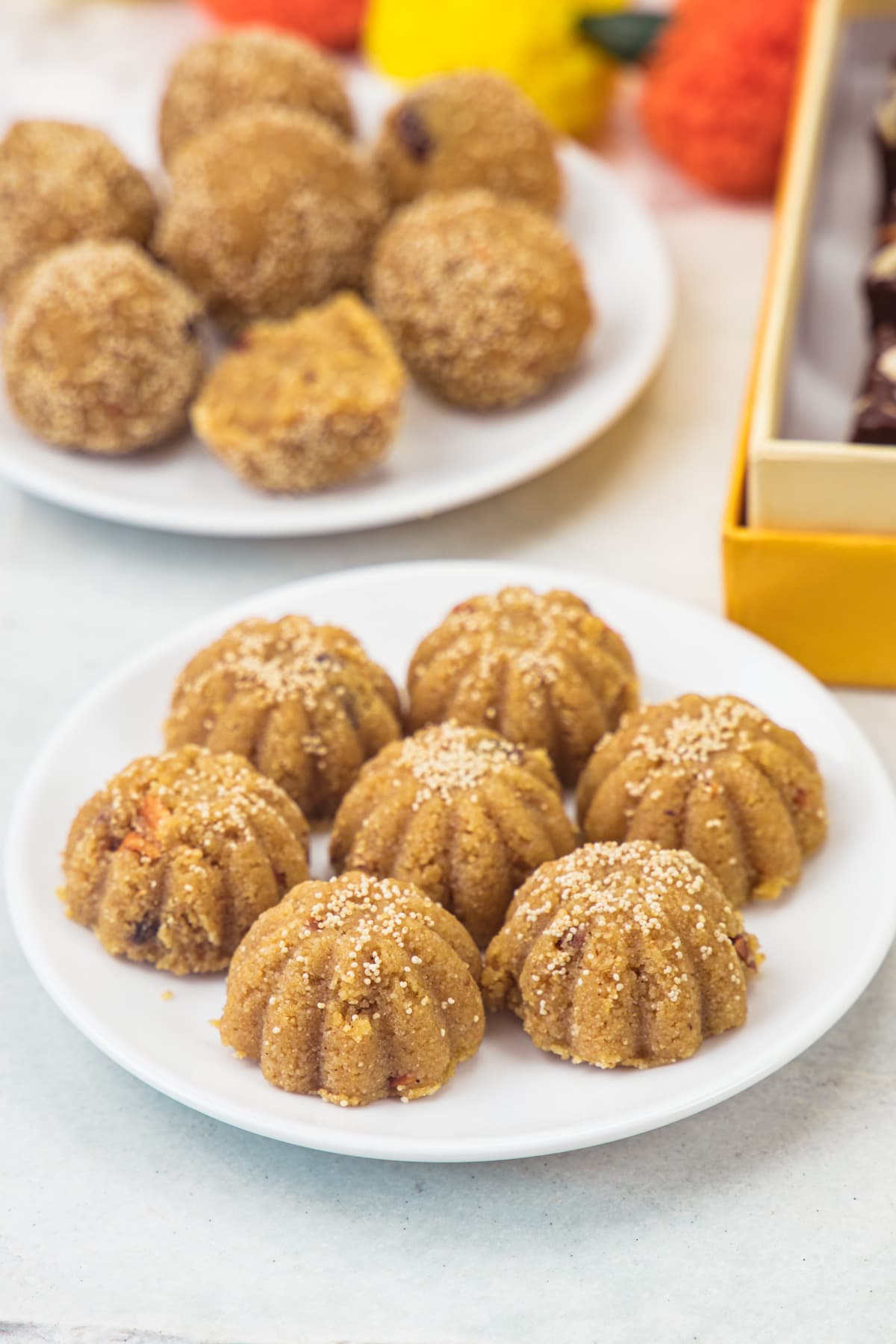 2 plates of churma ladoo with box and flowers in the back.