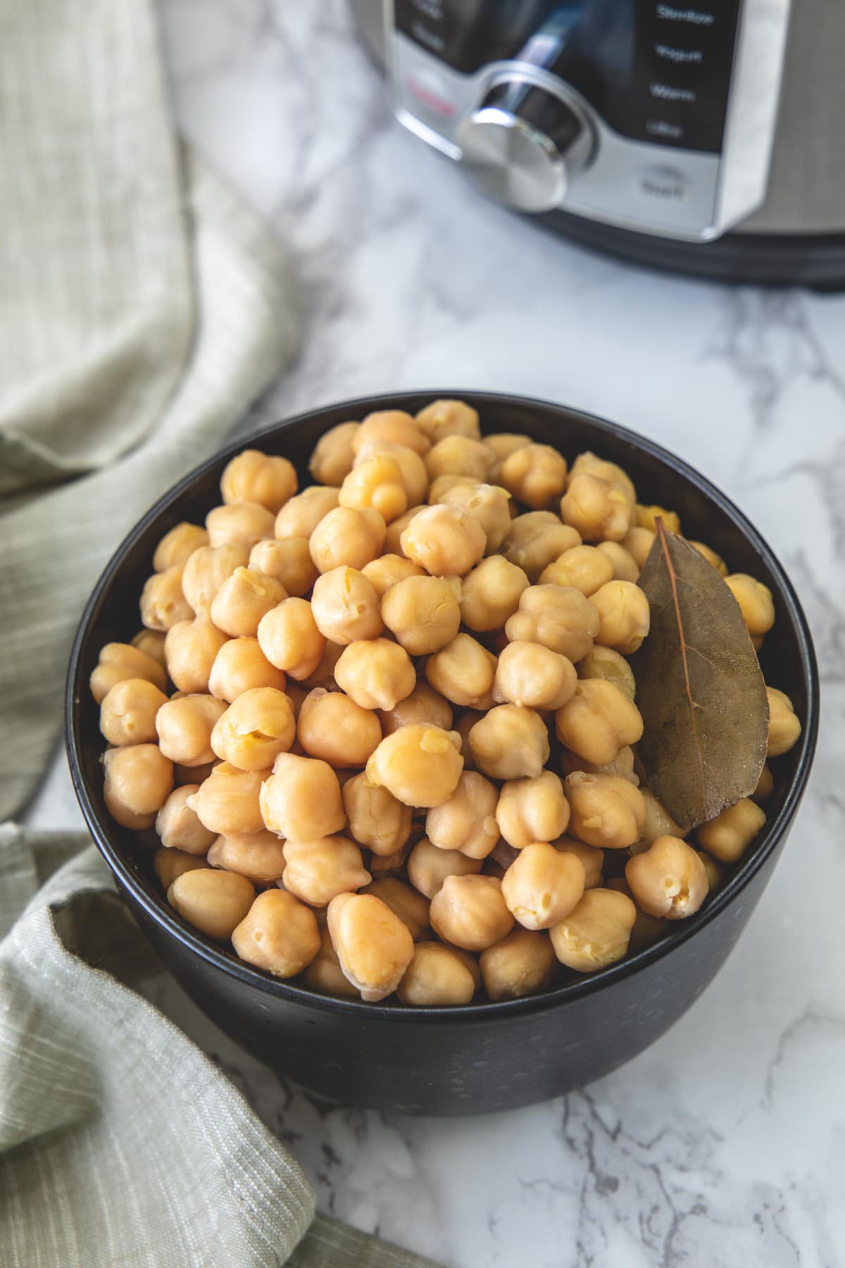 Instant pot cooked chickpeas in a bowl with a bay leaf, napkin on side and instant pot in back.