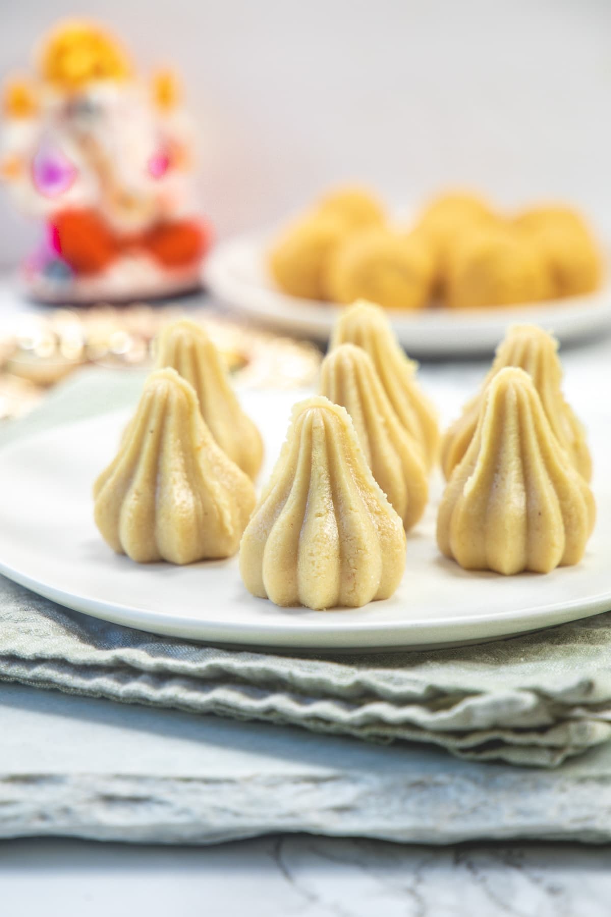 kaju modak in a plate with a plate of laddo and Ganesh idol in the back.