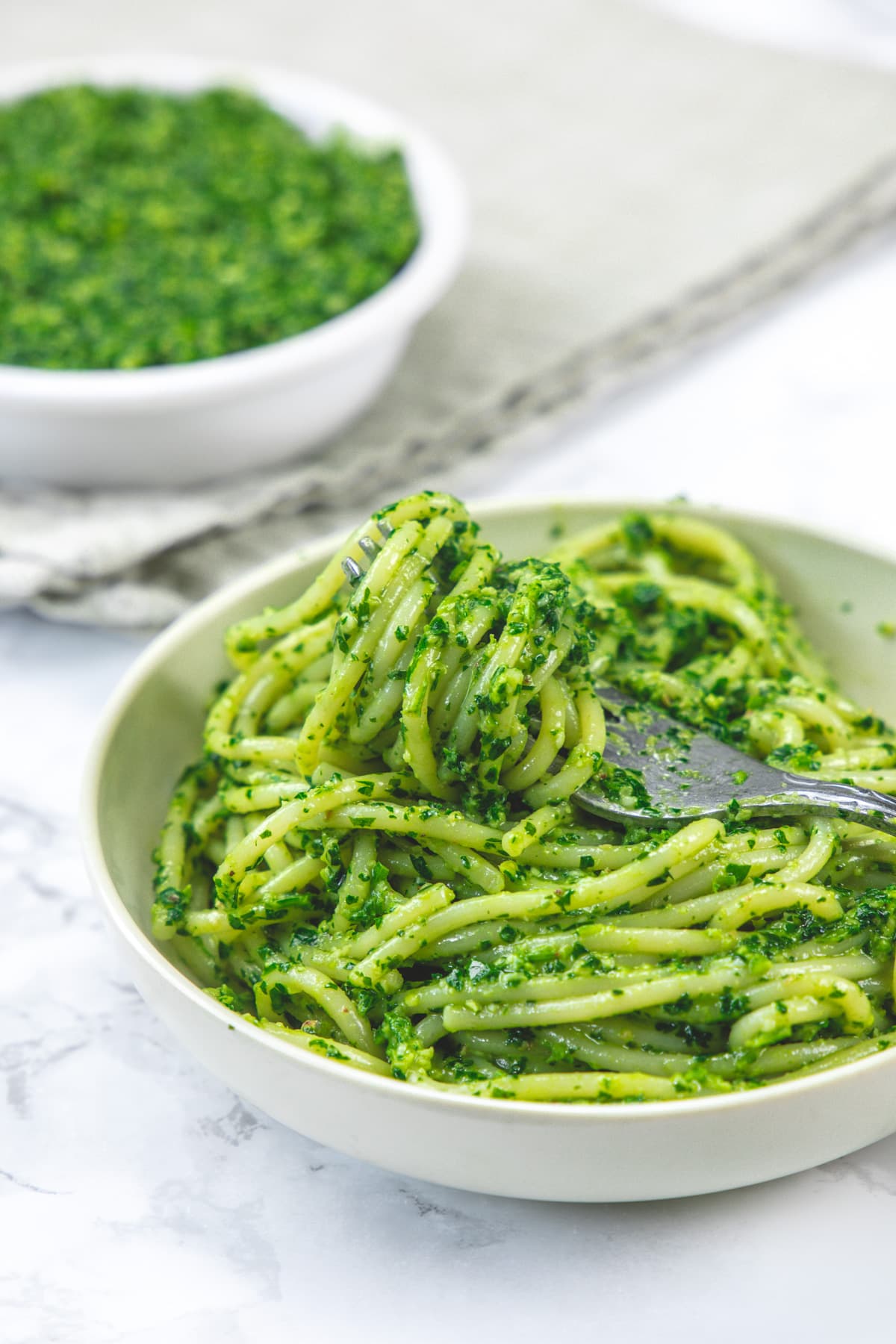 Pesto spaghetti pasta taking with a fork and parsley pesto bowl in the back.