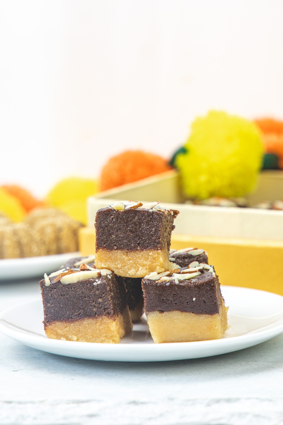 Stack of 3 chocolate burfi in a plate with flowers in the back.