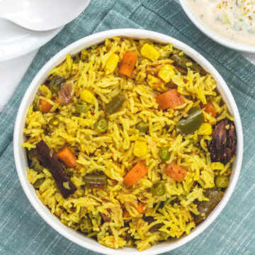 Veg pulao in a bowl with a bowl of raita and 2 spoons in the back.