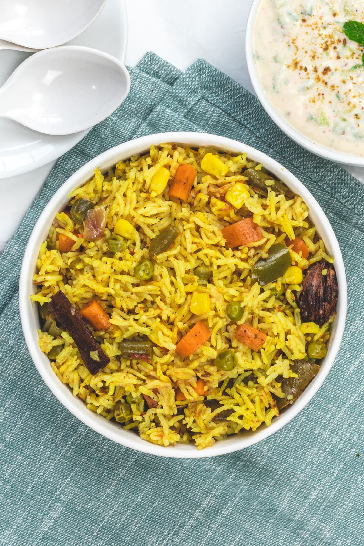 Veg pulao in a bowl with a bowl of raita and 2 spoons in the back.