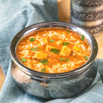 Paneer lababdar in a steel serving wil with glass in the back.