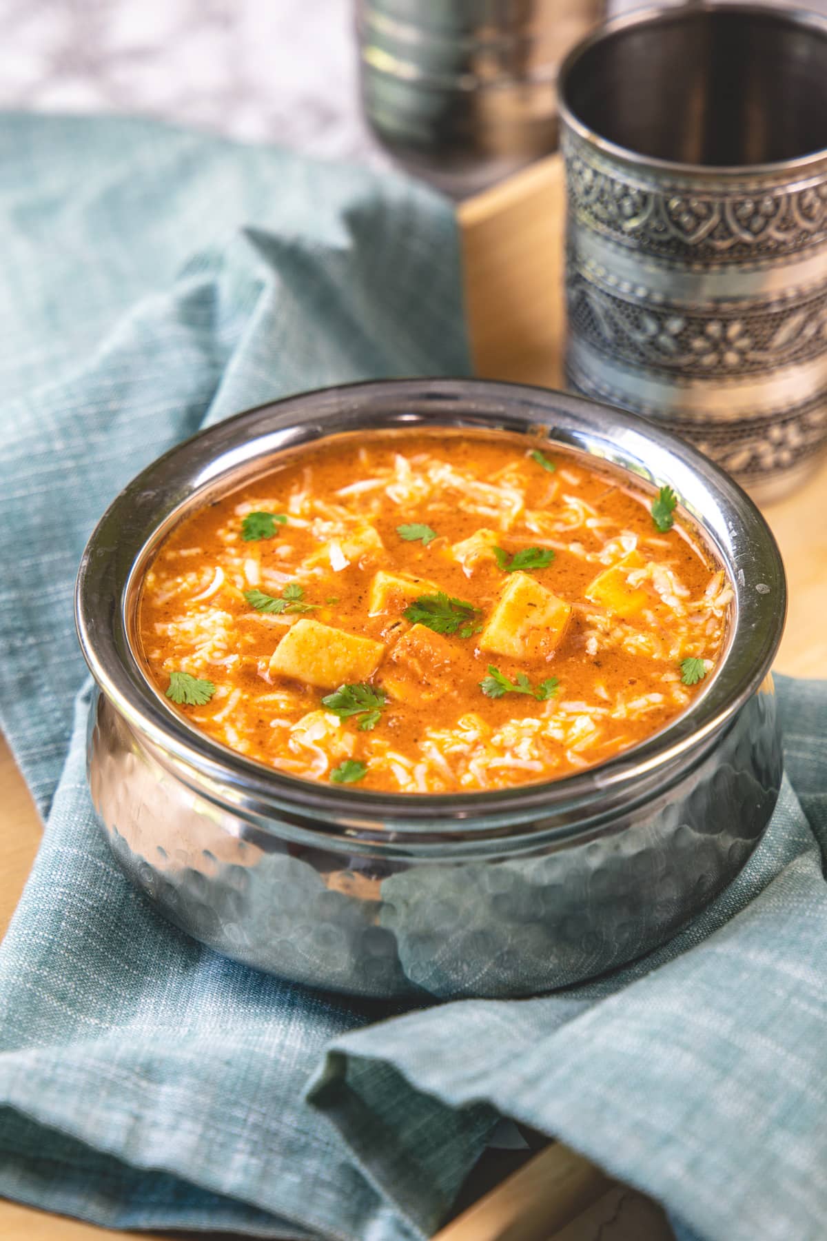 Paneer lababdar in a steel serving wil with glass in the back.