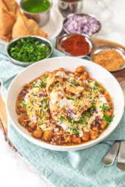 A plate of samosa chaat with more samosa, onion, chutney, cilantro in the back.