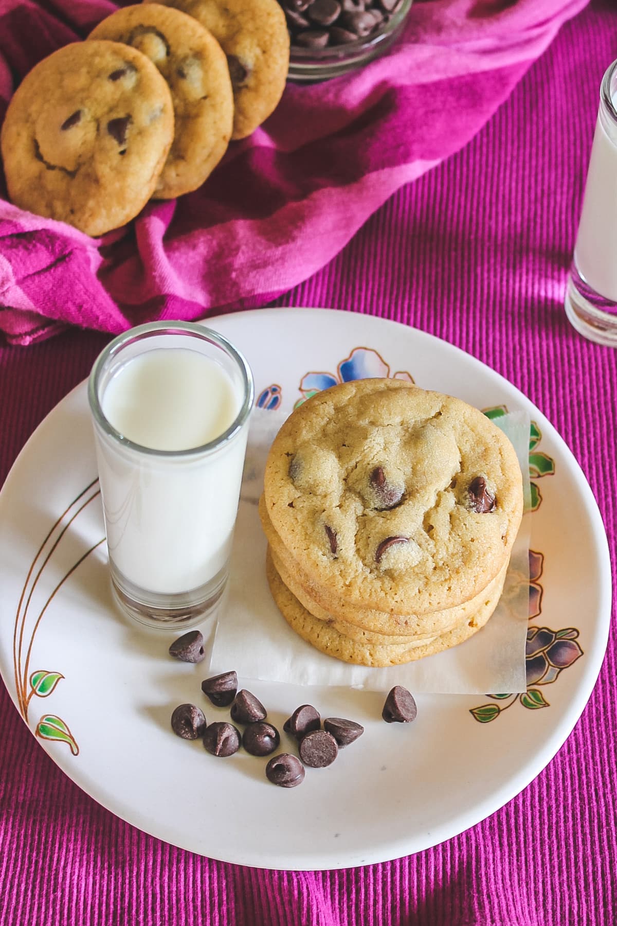 Chocolate chip cookies in a plate with a few chocolate chips and a shot glass with milk.