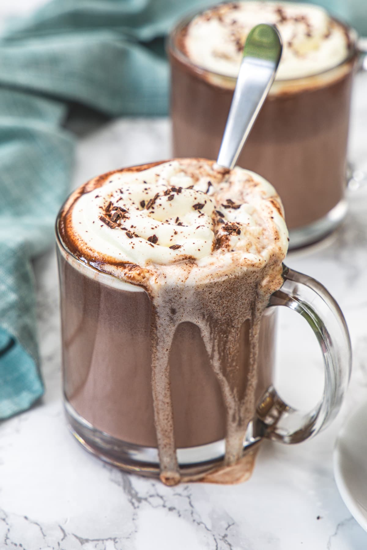 A mug of hot chocolate with a spoon into the mug and some hot chocolate is spilling.