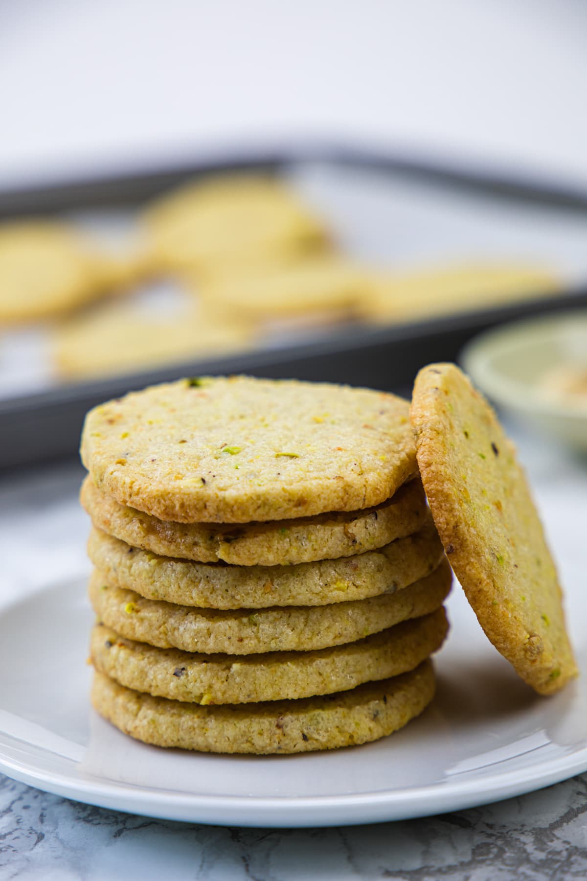 A stack of lemon pistachio cookies in a plate with another cookie is leaning on the stack.