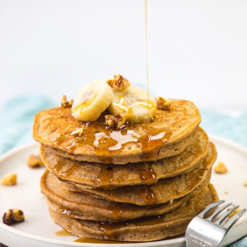 A stack of eggless banana pancakes topped with banana, nuts and drizzling syrup.