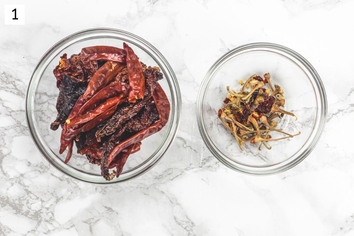 Two types of dried red chilies in a bowl and seeds, stems removed in another bowl.