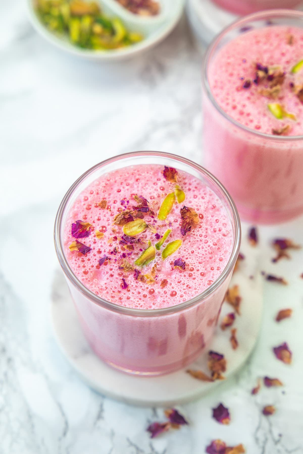 2 glasses of rose lassi garnished with pistachios and rose petals.