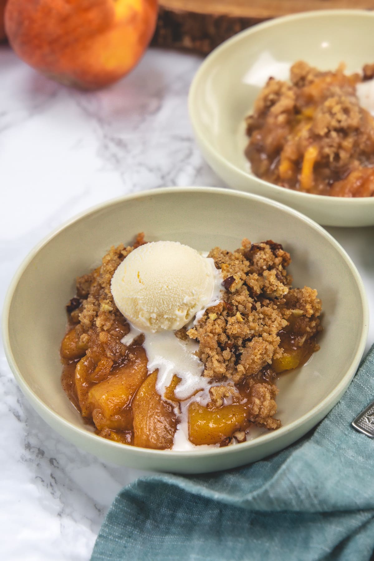 2 plates of peach crumble with vanilla ice cream and peaches in the back.
