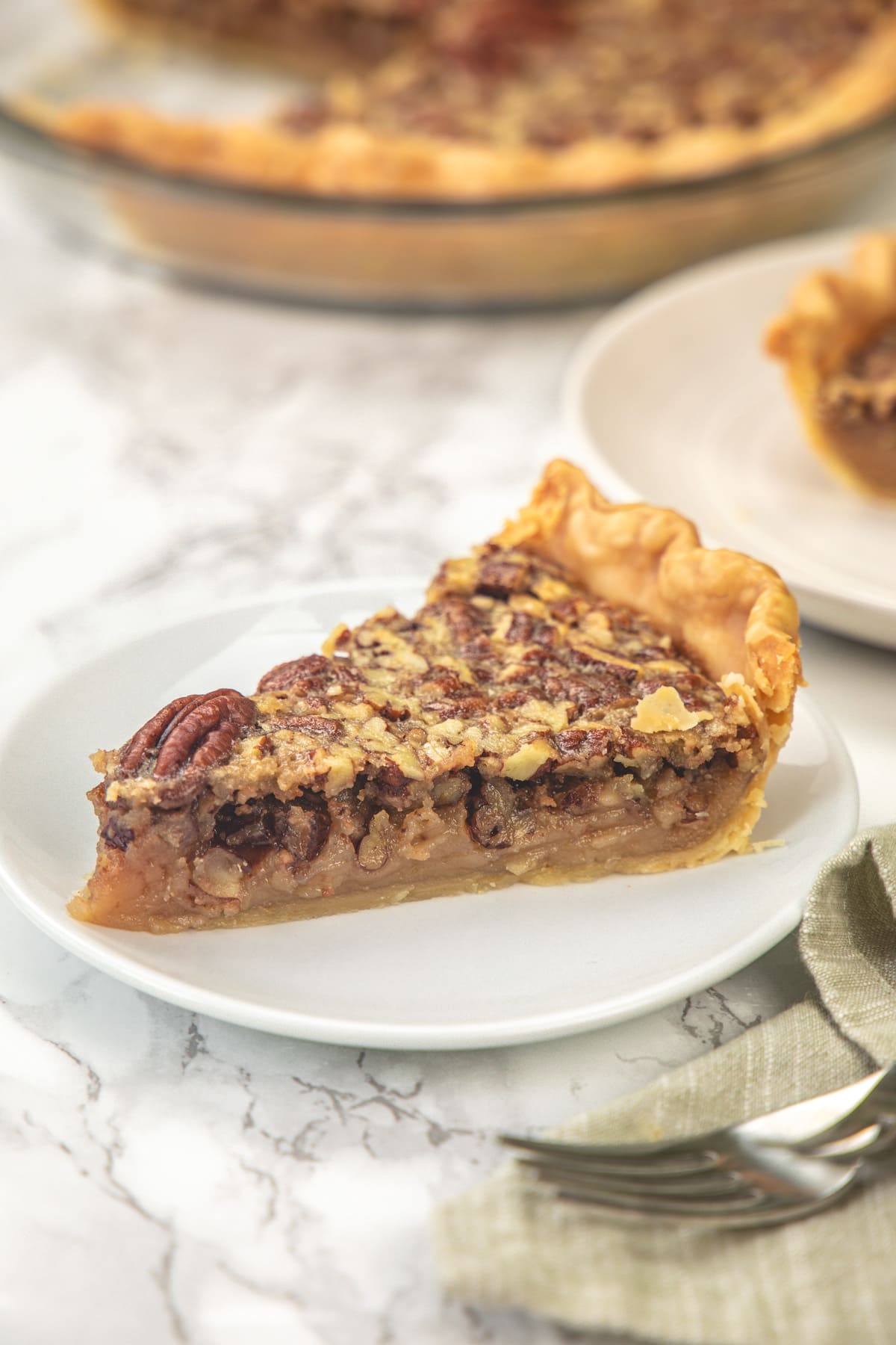 A slice of eggless pecan pie in a plate with fork and pie plate in the back.