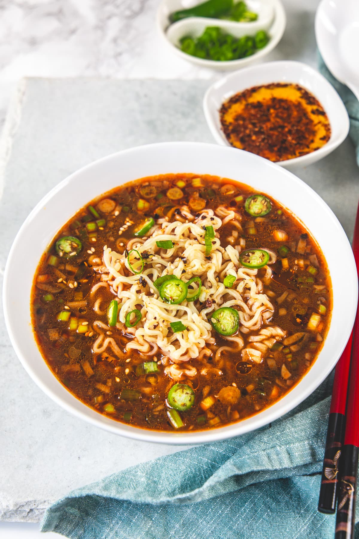 Spicy ramen noodles served in a bowl with a sides of chili oil and sliced green chilies.