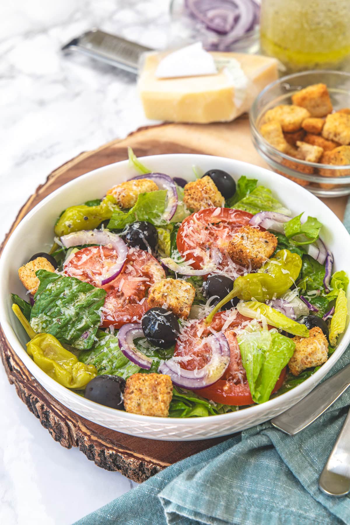 A bowl of copycat Olive Garden salad with block of Parmesan cheese and croutons in the back.